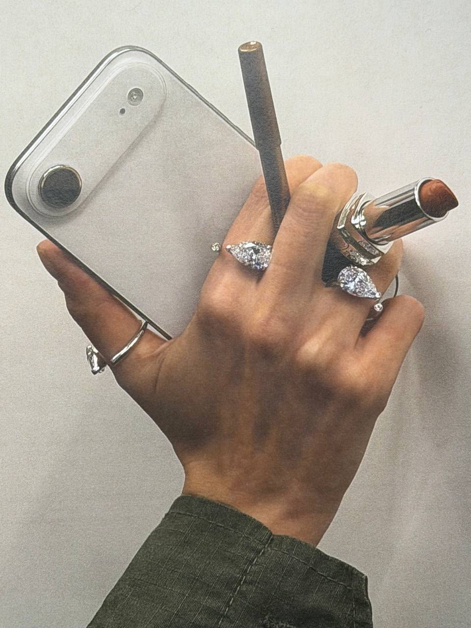 Hand wearing diamond rings while holding a phone, lipstick and pencil